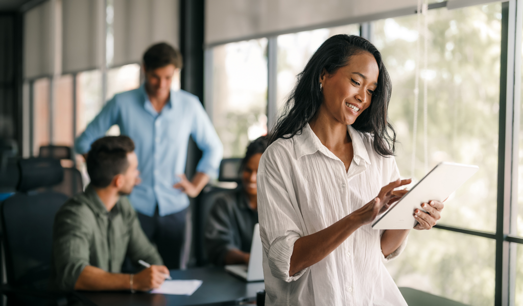 Woman reviewing a tablet in an office with colleagues in the background, representing a sales enablement team at work